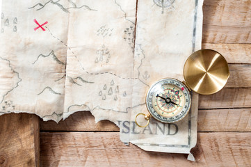Nautical compass with fake treasure map of abstract island with red cross on wooden table