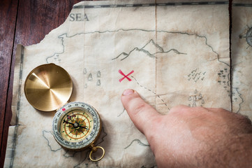 Man point with finger into red cross on Ancient Treasure fake map with gold compass on wooden desk