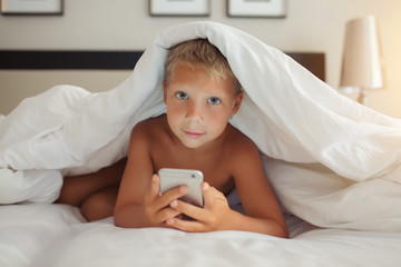 Obraz na płótnie Canvas A beautiful white boy lies in bed under a blanket and holds a gadget in his hands.