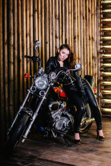 Obraz na płótnie Canvas Young sexy fashion woman in leather jacket and leather pants sitting on a motorcycle in Studio on a background of wooden wall