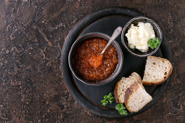 Bowl of red caviar with spoon served with sliced bread, butter and herbs on black wooden chopping board over brown texture background. Top view with space