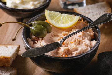 Black bowl of salmon pate with red caviar served with butter, sliced bread, capers, vintage knife and herbs on wooden serving board with textile linen over brown texture background. Close up