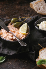 Black bowl of salmon pate with red caviar served with butter, sliced bread, capers, vintage knife and herbs on textile linen napkin over brown texture background. Close up