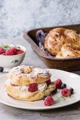 Filling and empty homemade choux pastry cake Paris Brest with raspberries, almond, sugar powder, rosemary on plate and oven tray with berries over gray texture background. French dessert.