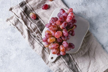 Bunch of red pink ripe wet grapes on white ceramic chopping board with linen kitchen towel over gray blue texture background. Top view with space