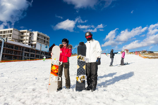 Young couple in ski suits, helmets and ski goggles standing with snowboards in a ski-resort