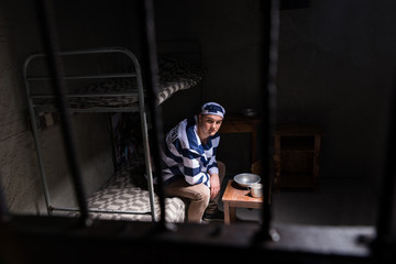 Fototapeta na wymiar View through iron door with prison bars on male wearing prison uniform sitting on a bed