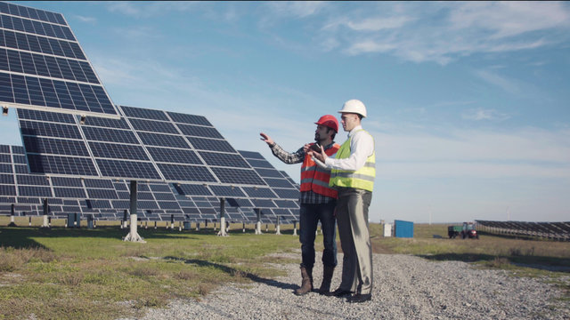 Two engineers in uniform and helmets talking at solar power station and using tablet.