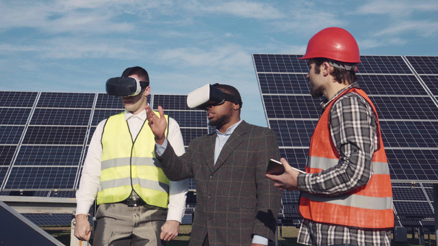 Workman and investor wear virtual reality glasses as they stand near array of solar panels