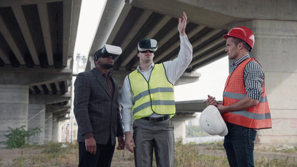 4K shot of business team of two architects and afro american foreman in suit uses virtual reality glasses to discuss a project of development while standing under overpass construction.