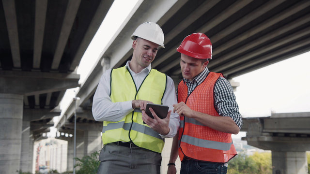 4K shot of Two young workers in red and yellow vests and hard hats lookig at tablet computer and discusses a project while standing under the bridge construction. Movement stabilized