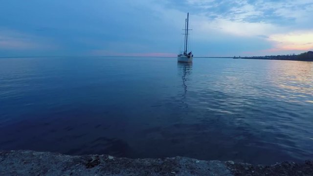 Two-masted white yacht approaches the concrete pier. View from the pier. Cloudy, evening, calm.