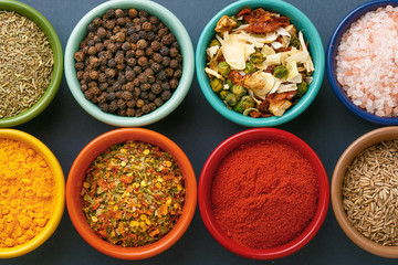 Obraz na płótnie Canvas Spices in colorful bowls viewed from above. Various seasonings on a dark background. Italian mix, cumin, chili pepper, curry powder, Himalayan salt, pepper, garlic, cinnamon, dried tomato. Top view