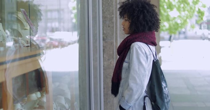 Side view of black woman with curly hair in casual clothing standing near shop window and looking ar jewelry.