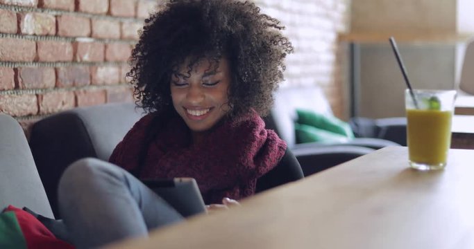 Charming young woman in casual outfit sitting at table with glass of fresh drink on it and browsing tablet smilingly.