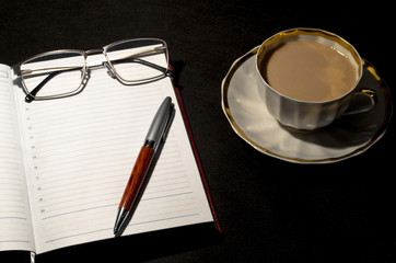 Open diary book with empty pages,white cofee cap, pencil and glasses on black background, close up. Business or education concept.