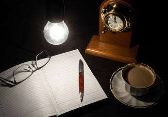Open diary book with empty pages, white cofee cap, pencil,glasses and wooden clock on black background under lights of switched on bulb. Business or education concept.