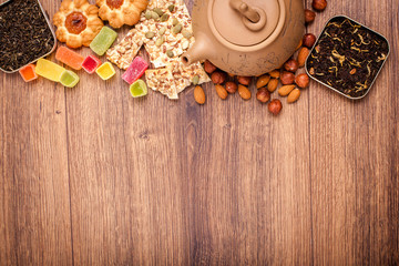 The composition of the tea on a wooden surface. brown teapot from clay with a scattering of tea, almond and hazelnut. Green, black tea. Cookies and gummy candies. Top view with copy space.