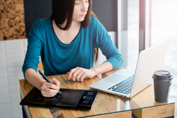 Young female freelance designer working on the laptop and graphic tablet while sitting at the cafe