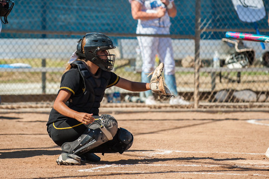 Softball catcher in black uniform reaching out with glove for the pitch.   