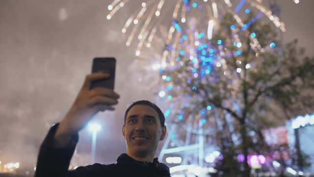Closeup of young cheerful man watching and making selfie picture with fireworks on smartphone camera outdoors