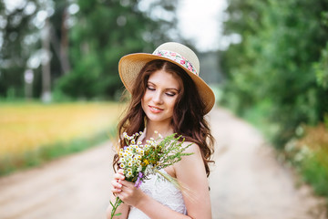 Fototapeta premium pretty woman beautiful young happy with long dark hair in white dress and hat holding small bouquet of flowers, nature background outdoors