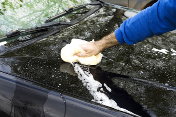 Dłoń mężczyzny myje maskę samochodu gąbką z dużą ilością piany.