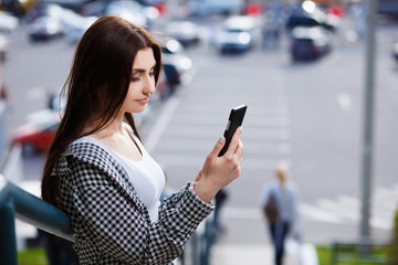 Young pretty stylish smiling woman reading message on smart phone in the street