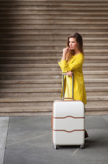 Young woman, dressed in yellow suit,  leans on the suitcase and looking at something