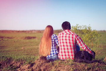 Young couple in plaid shirts and shorts and the dog is sit back and admire the scenery. Tourists rest on the hill in front of the grass field. Toned
