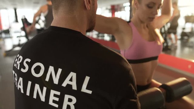 Fit female doing rear lat pull-downs under supervision of her personal trainer