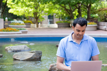 Closeup portrait, young man in blue polo typing on laptop with background of pond and trees