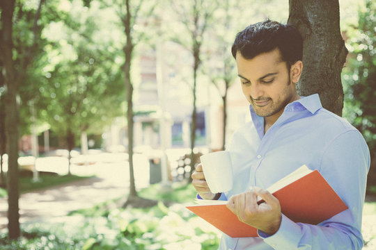 Closeup portrait young business man reading red book and drinking from mug, standing outside on sunny day, isolated green trees background