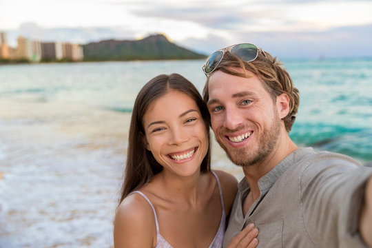 Selfie couple on Waikiki beach taking pictures with smartphone during night out walk on beach summer vacations in Honolulu, Hawaii. Travel destination. Young people having fun on hawaiian holidays.