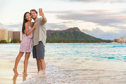 Couple walking on beach at sunset taking selfie picture on mobile phone relaxing together on Waikiki beach, Honolulu, Hawaii travel vacation. Romantic holiday destination for honeymoon.