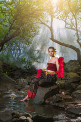 Thai woman in tradition silk dress sit on the rock in the forest. with light ray and fog