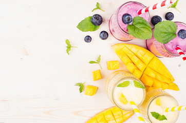Yellow and violet  fruit smoothie in glass jars with straw, mint leaves, mango slices and berry, top view. Soft white wooden board background, copy space.