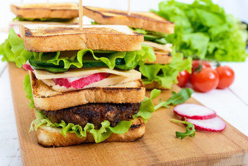 Multilayered sandwiches with a juicy cutlet, cheese, radish, cucumber, lettuce, arugula on a cutting board on a white wooden background.