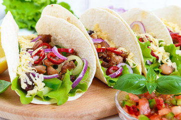 Tacos is a traditional Mexican dish. Tortilla stuffed with chicken, bell and hot peppers, beans, lettuce, cheese, blue onion with salsa sauce on white wooden background.