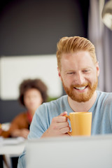 Male using laptop in modern office while sitting and drinking coffee. colleague in background