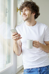 Starting the day with coffee and reading messages. Shot of a young man having a cup of coffee and text messaging while looking out his apartment window.