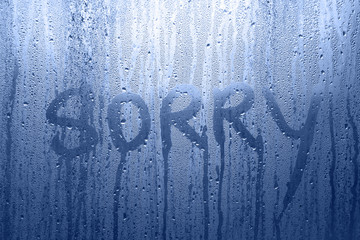 Conceptual sorry word handwritten message on the rainy glass window background. Blue color tone...