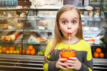 A girl stands near a showcase and drinks juice Through the straw in a cafe