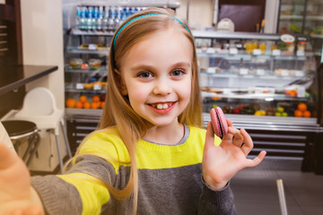 A little girl stands near a showcase with macaroon and makes selfie