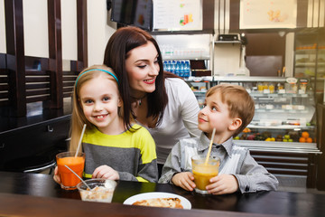 young Mom with her chidren is sitting in a cafe and drinking juice