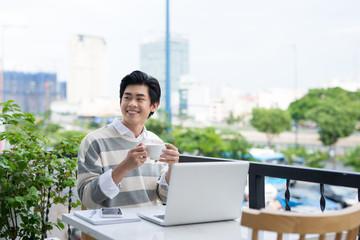 Handsome asian young man working on laptop and smiling while enjoying coffee in cafe