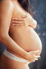 Fototapeta na wymiar Pregnant woman with a slender beautiful body hugging her belly and chest standing on a gray background
