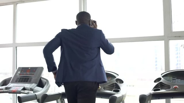 Busy African-American male in suit running on treadmill and talking on the phone