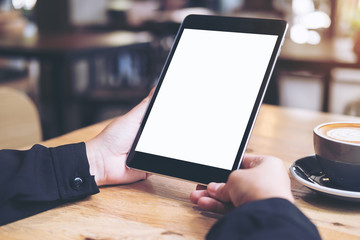 Mock up image of business woman's hands holding black tablet with white blank screen and coffee cup on wooden table in cafe