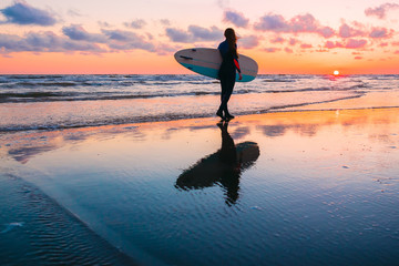 Young and sporty girl go to surfing. Beautiful woman in wetsuit and sunset on ocean